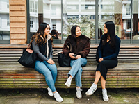 three women sit outside on a wooden bench talking