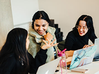 three students sit talking at a desk with laptops in front of them